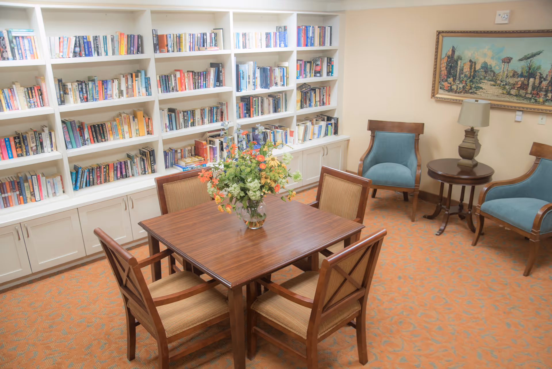 A cozy reading room with a wooden table surrounded by four chairs, a vase with flowers on the table, a large white bookshelf filled with books along the wall, two blue upholstered armchairs with a round wooden side table and lamp between them, and a framed artwork hanging on the beige wall.