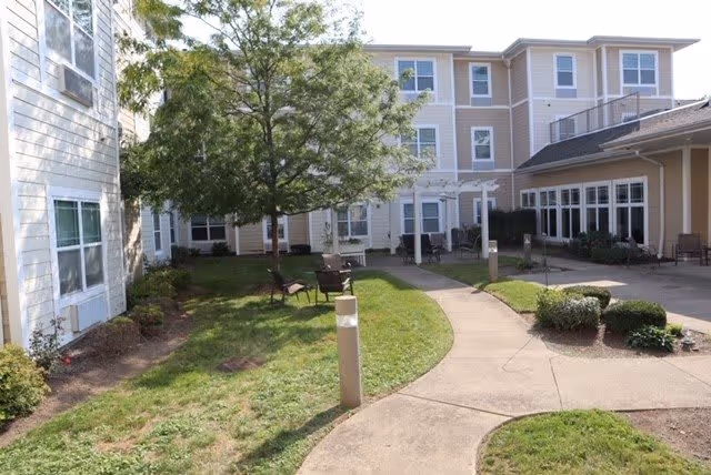Outdoor courtyard area of a senior living facility with a paved walkway, green grass, bushes, and a tree. The building surrounds the courtyard with multiple windows and a covered patio area with chairs.