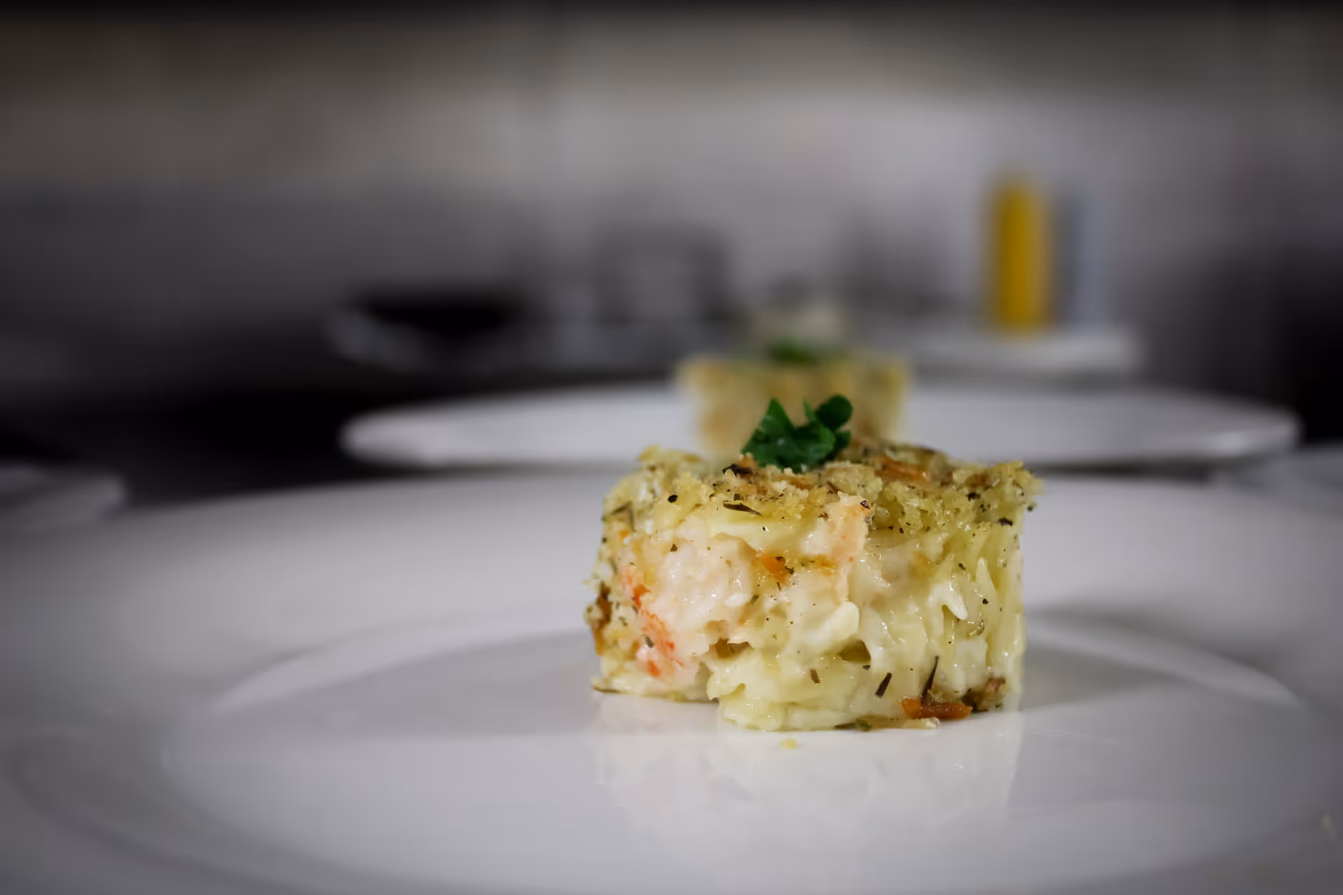 Close-up of a small, round serving of creamy baked rice dish garnished with herbs on a white plate, with another similar serving blurred in the background.