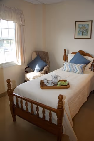 A bright single-occupancy bedroom with a wooden bed topped with pillows and a tea tray, an armchair by a window, and framed artwork on the wall.