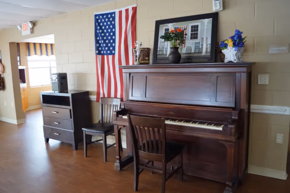 Upright piano with chairs, an American flag, and decorative items in a communal living room area.