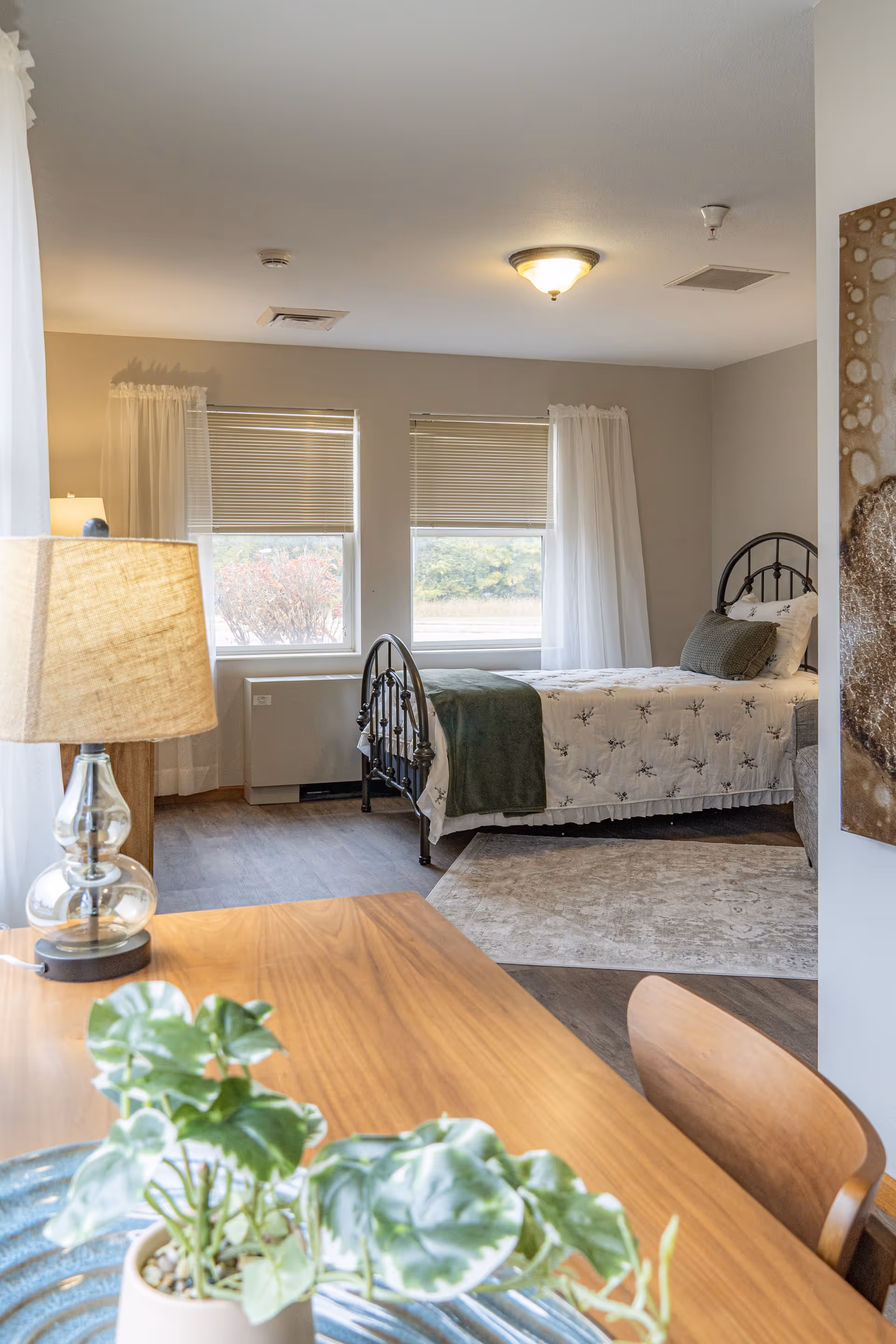 A bright and cozy bedroom with two windows covered by blinds and sheer white curtains. The room features a single bed with a metal frame, white bedding with a floral pattern, and a green throw blanket and pillow. In the foreground, there is a wooden table with a potted plant and a glass lamp with a beige lampshade. The floor is wood with a light-colored area rug near the bed.