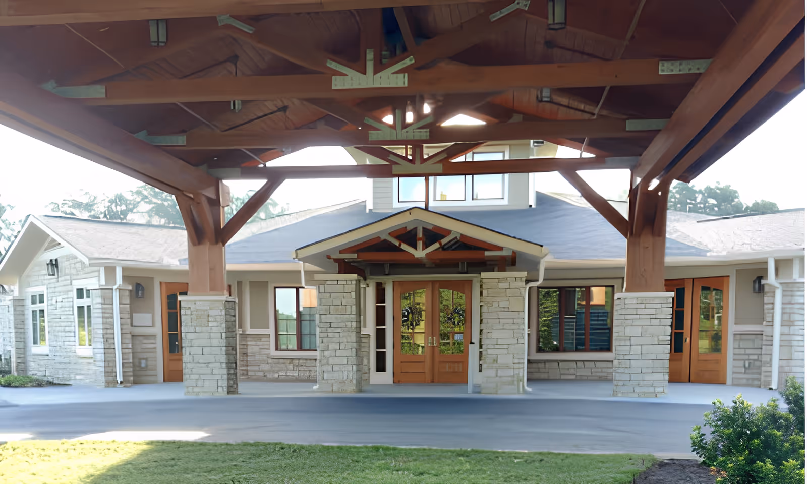 Front entrance of a senior living facility with a covered driveway supported by wooden beams and stone pillars. The building features large windows and wooden double doors, with a well-maintained lawn and bushes in the foreground.