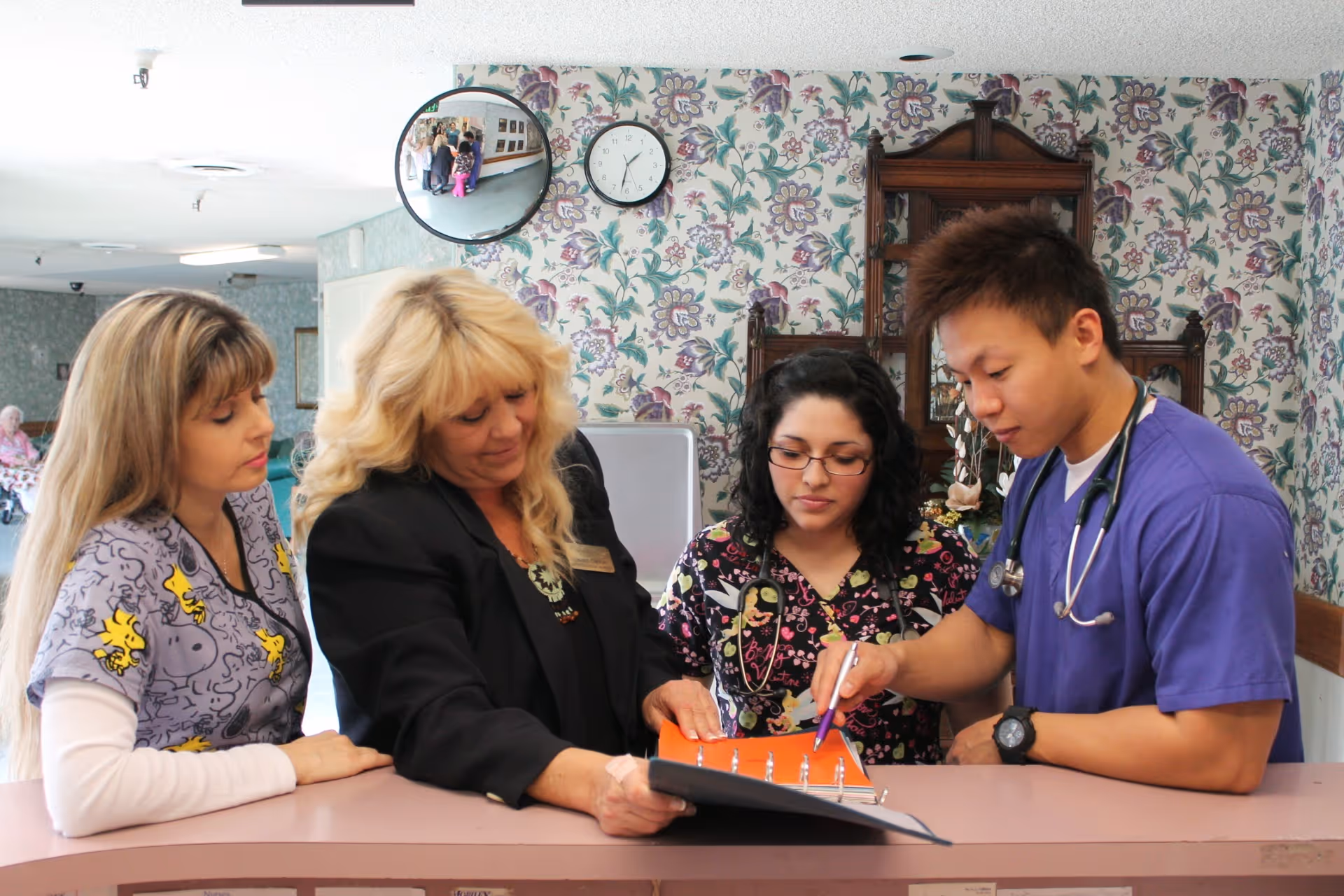 Four healthcare workers standing behind a reception desk in a nursing facility. They are looking at and discussing a binder with documents. The background features floral wallpaper, a clock, and a convex mirror reflecting part of the room.