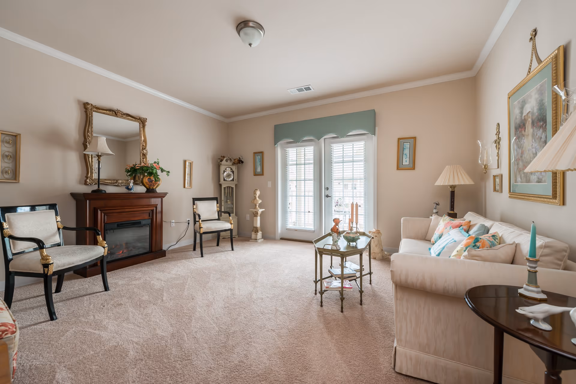 A cozy living room with beige walls and carpet, featuring a cream-colored sofa with colorful pillows, two black and gold armchairs, a wooden electric fireplace with a decorative mirror above it, a glass coffee table with decorative items, and a grandfather clock near French doors with white blinds leading outside.