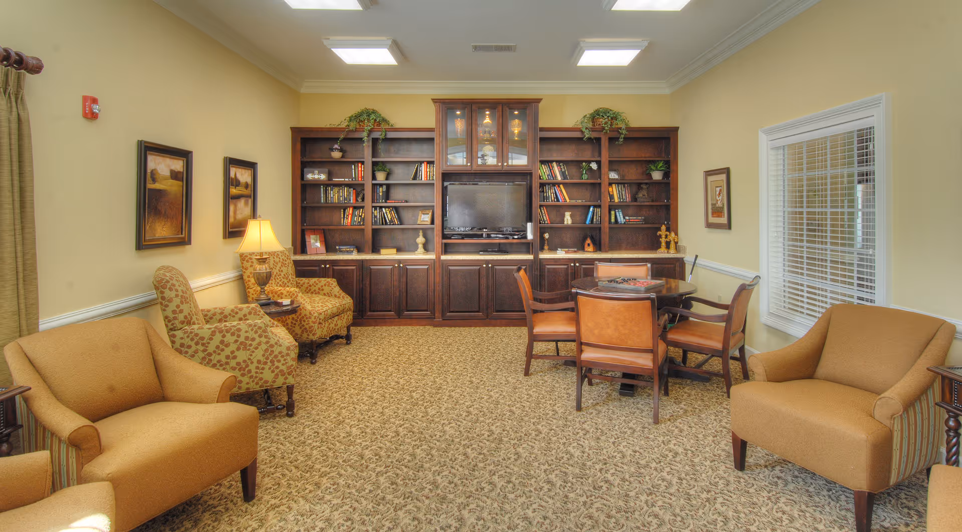 A cozy living room with beige walls and carpet, featuring a dark wooden built-in bookshelf and cabinet unit with a TV in the center. The room has several armchairs and a round table with four chairs. There are framed paintings on the walls, a table lamp, and a window with white blinds.