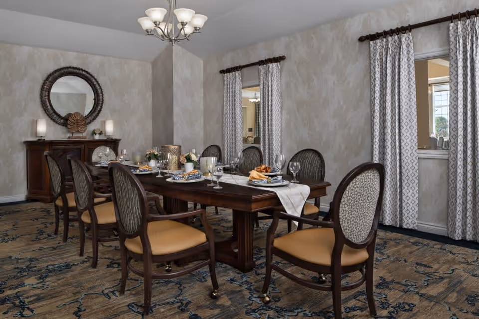 A formal dining room with a long wooden table set for a meal, surrounded by eight upholstered chairs. The table is decorated with candles, flowers, and neatly folded napkins. The room features patterned curtains, a large round mirror above a wooden sideboard, and a chandelier hanging from the ceiling.