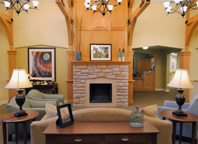 A cozy living room area featuring a stone fireplace with a wooden mantel, flanked by two armchairs and side tables with lamps. Above the fireplace is a framed picture, and decorative vases are placed on the mantel. The room has warm lighting from ceiling chandeliers and wooden architectural beams. In the background, there is a hallway leading to other rooms.