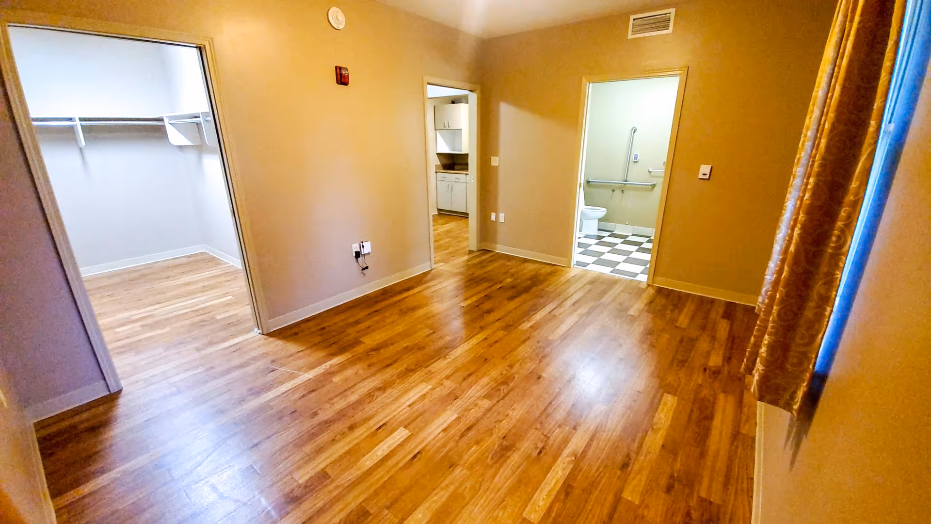 Empty room with wooden flooring and beige walls, featuring an open doorway to a walk-in closet on the left, a doorway to a kitchen area in the center, and a doorway to a bathroom with checkered black and white floor tiles and a toilet on the right. A window with patterned curtains is visible on the right side.