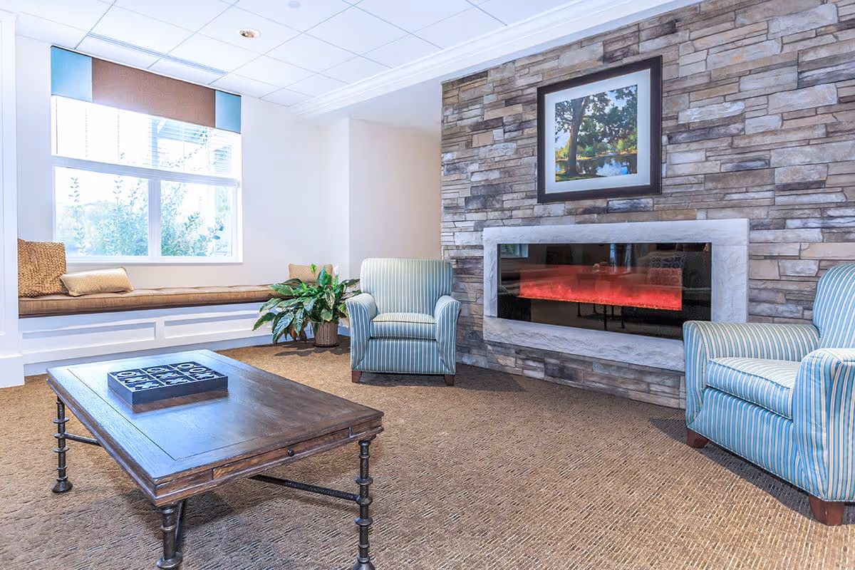 Bright sitting area with two striped armchairs, a low wooden coffee table, a window bench, and a stone-wall electric fireplace.