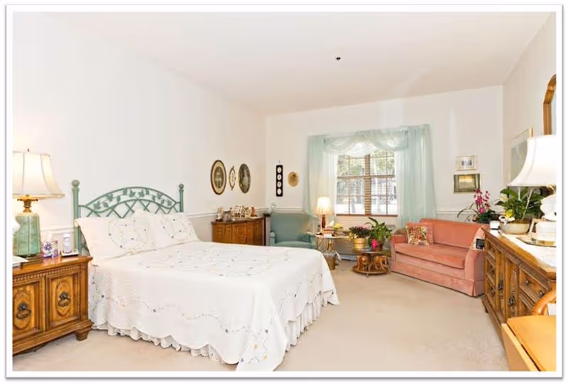 A bright and cozy bedroom in a senior living community featuring a bed with a decorative green headboard and white bedding, wooden nightstands with lamps on either side, a wooden dresser, a pink sofa, a green armchair, a window with light green curtains, and various decorative items and plants.