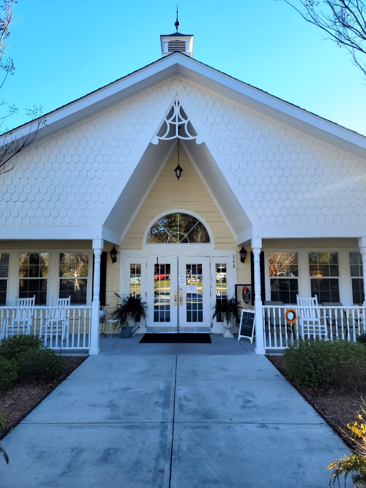 Front entrance of a building with a peaked roof and decorative white trim. The building has double glass doors with white frames, flanked by windows on both sides. There are white rocking chairs on the porch and potted plants near the entrance. A concrete walkway leads up to the door, and there is a small blackboard sign on the right side of the porch.