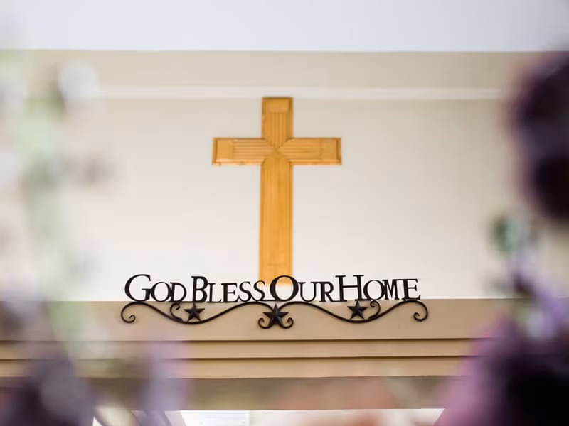 A wooden cross mounted on a beige wall above a decorative metal sign that reads 'God Bless Our Home' with star accents.