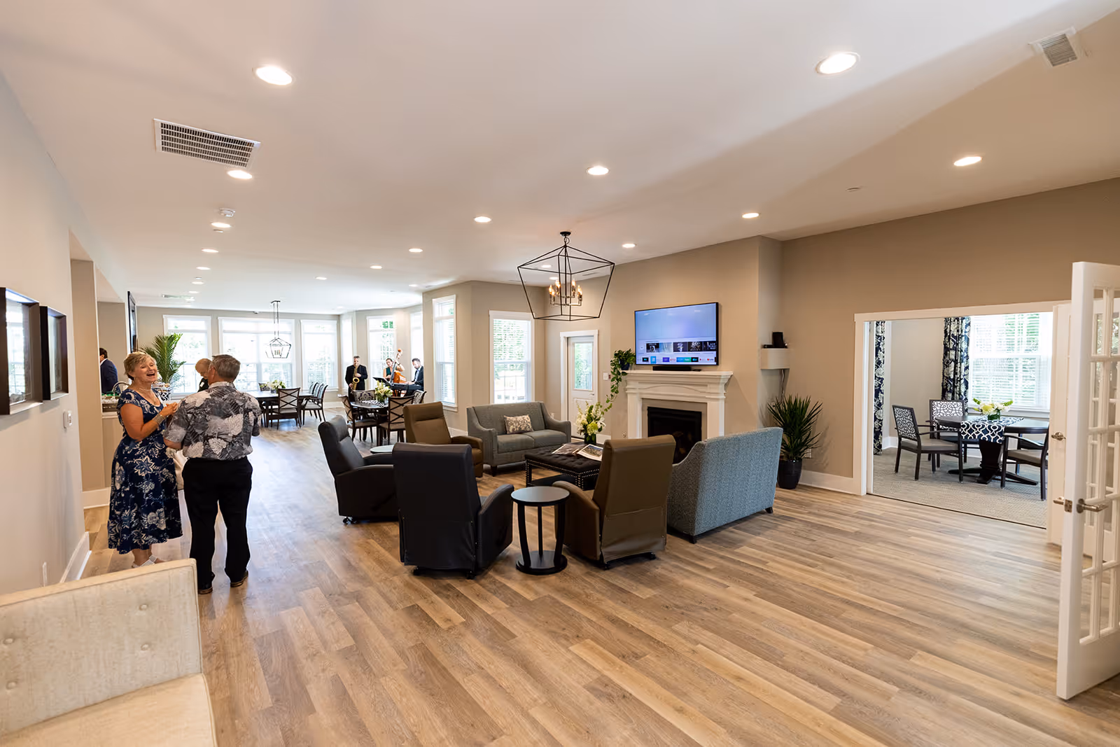 A spacious and well-lit living room area in a senior living facility with modern furniture including sofas and armchairs arranged around a coffee table and a fireplace with a mounted flat-screen TV above it. In the background, there are several people socializing near a dining area with tables and chairs, and a separate room with a table and chairs visible through an open doorway. The room features wood flooring, recessed lighting, and large windows allowing natural light.