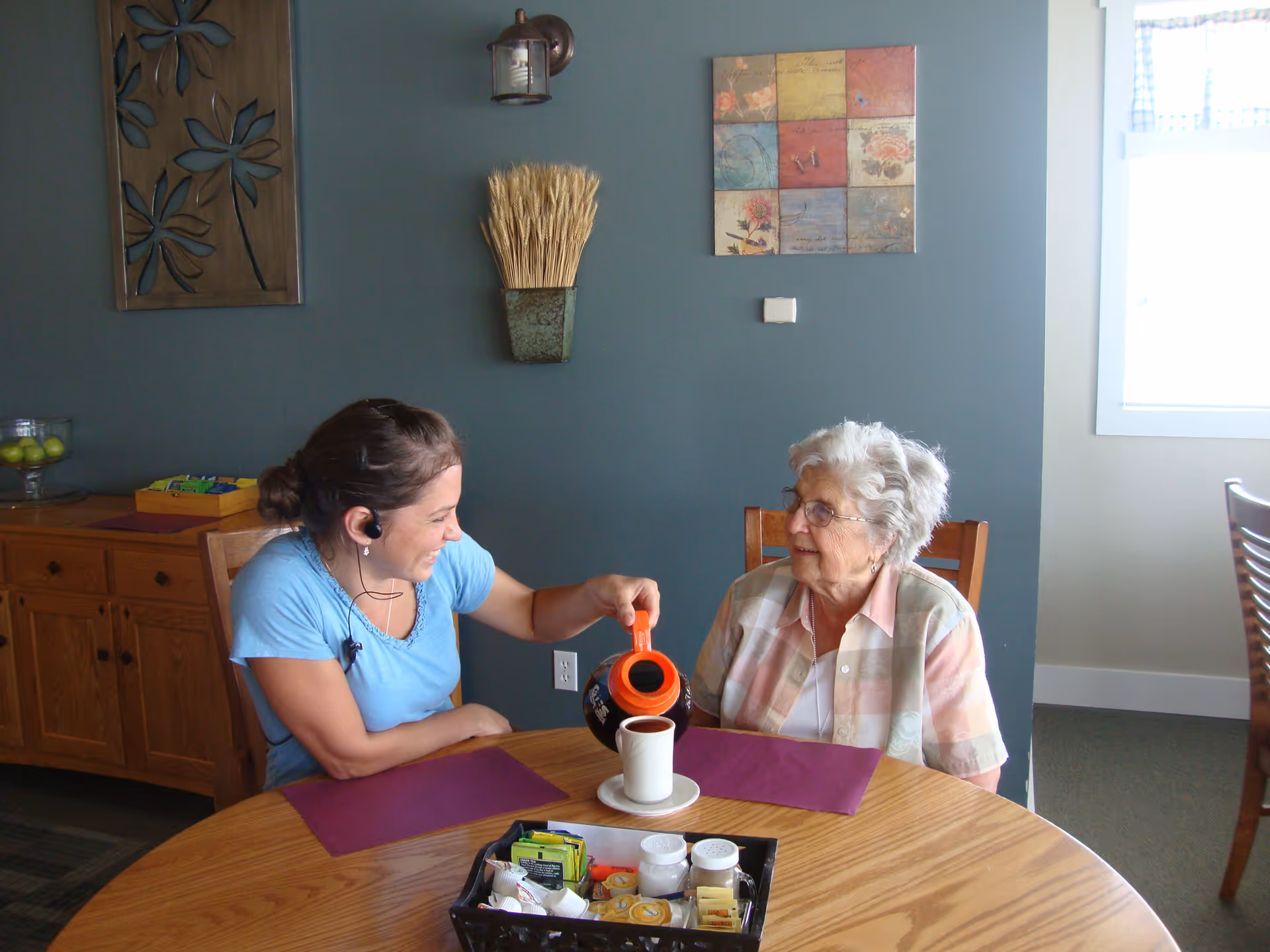 A caregiver pours coffee for an elderly woman seated at a dining table in a communal dining room.
