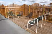 Outdoor patio area with wooden decking and a wooden privacy fence. The space includes several chairs and a small round table, with a clear roof overhead providing shelter.