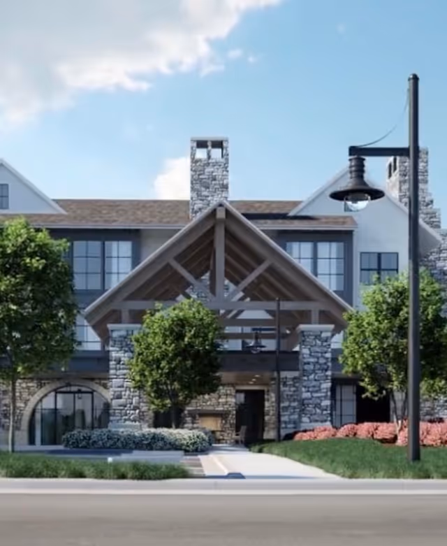 Front exterior view of a senior living facility named Village Park Milton, featuring a stone and wood facade with large windows, a covered entrance supported by stone pillars, landscaped greenery including trees and bushes, and a street lamp in the foreground under a partly cloudy sky.
