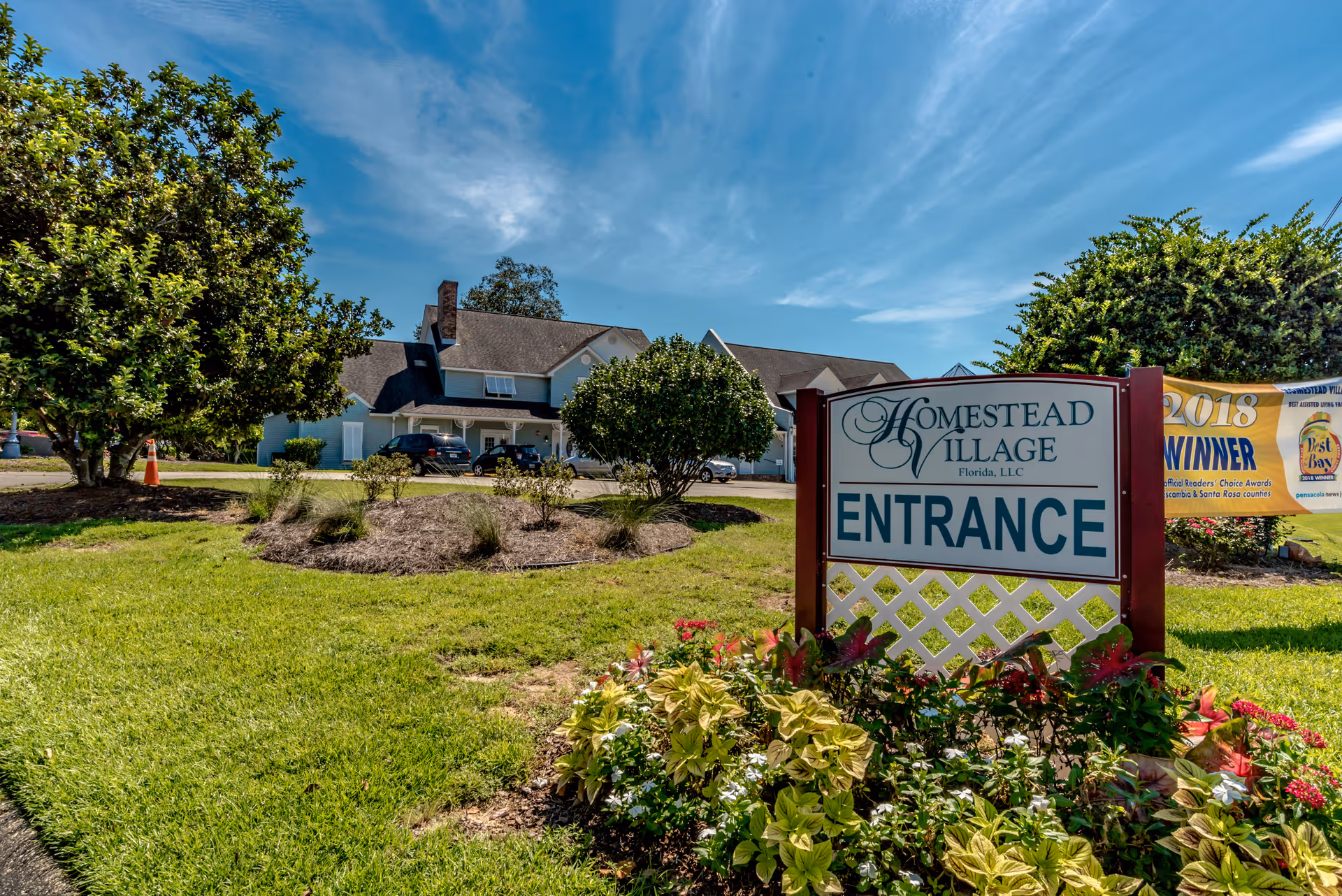 Entrance sign for Homestead Village in front of a landscaped lawn with the facility building and trees under a blue sky.