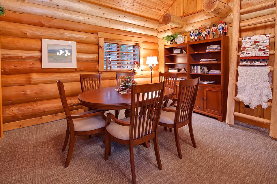 A cozy dining room with wooden log walls and ceiling, featuring a round wooden dining table surrounded by six cushioned chairs. A wooden bookshelf filled with books and decorative nutcracker figurines stands against the wall. A window with wooden blinds and a framed picture on the wall add to the warm ambiance. A floor lamp and a wooden rack holding a quilt and a crocheted blanket complete the room.