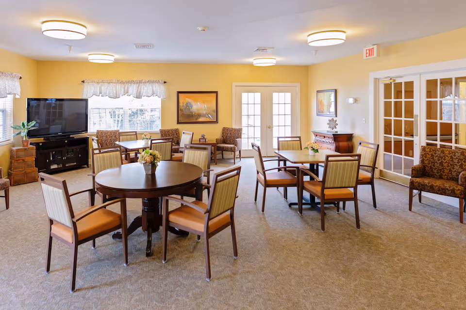 Well-lit communal dining/activity room with round and rectangular tables, chairs, a TV, and decorative furnishings.
