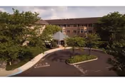 Exterior view of a multi-story senior living community building with a circular driveway and landscaped greenery including trees and shrubs under a partly cloudy sky.