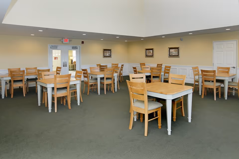 A spacious dining room with multiple wooden tables and chairs arranged neatly on a green carpet. The walls are painted light yellow with white wainscoting and decorated with framed pictures. Double doors with glass panels and an exit sign are visible at the back of the room.