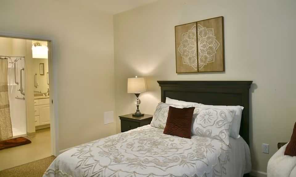 Cozy bedroom with a made bed, nightstand and lamp, wall art above the headboard, and an open doorway showing an adjacent bathroom.