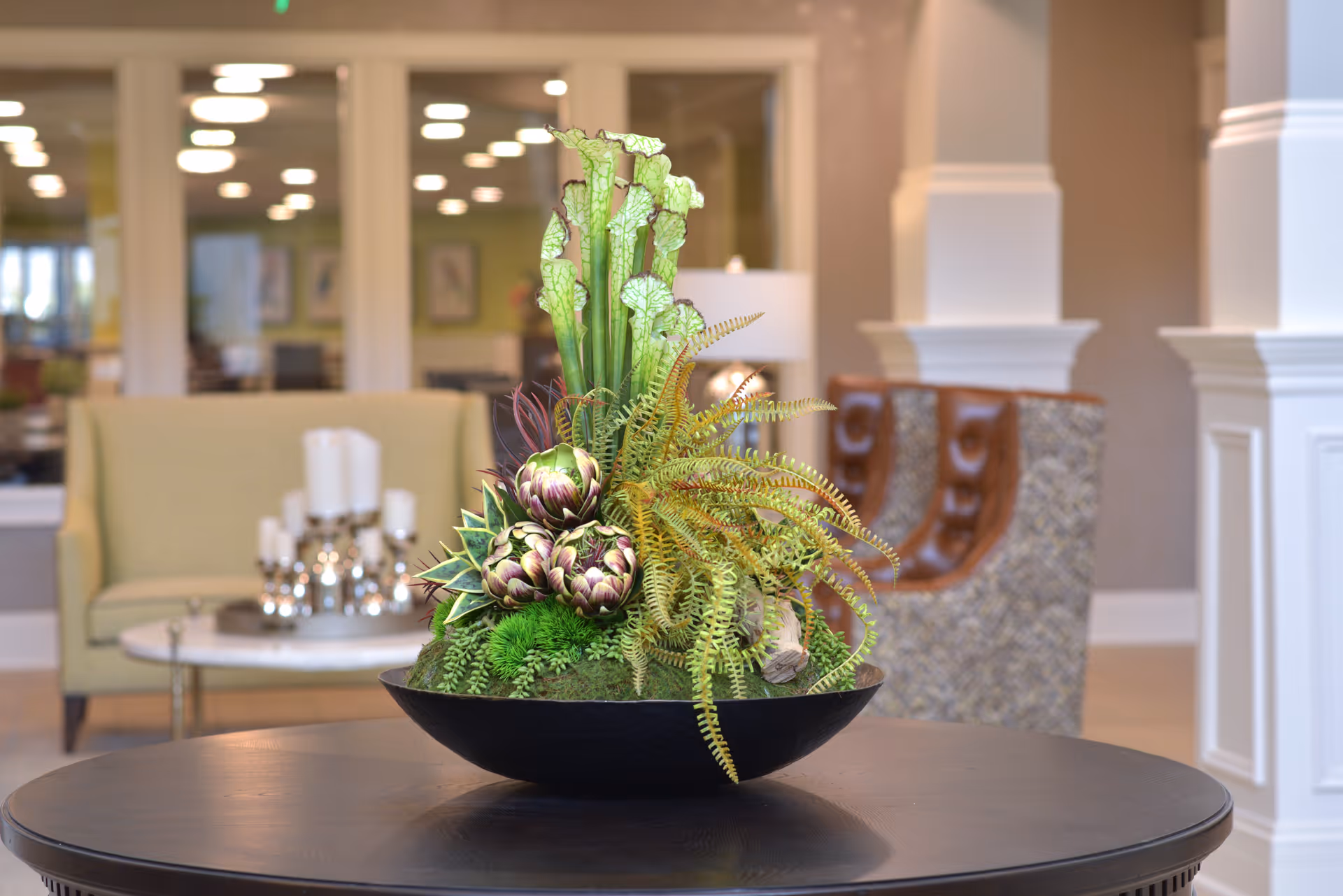 A decorative floral arrangement with green plants and artichoke-like flowers in a black bowl on a round wooden table, set in a well-lit living room area with beige and patterned chairs and a candle centerpiece on a side table in the background.