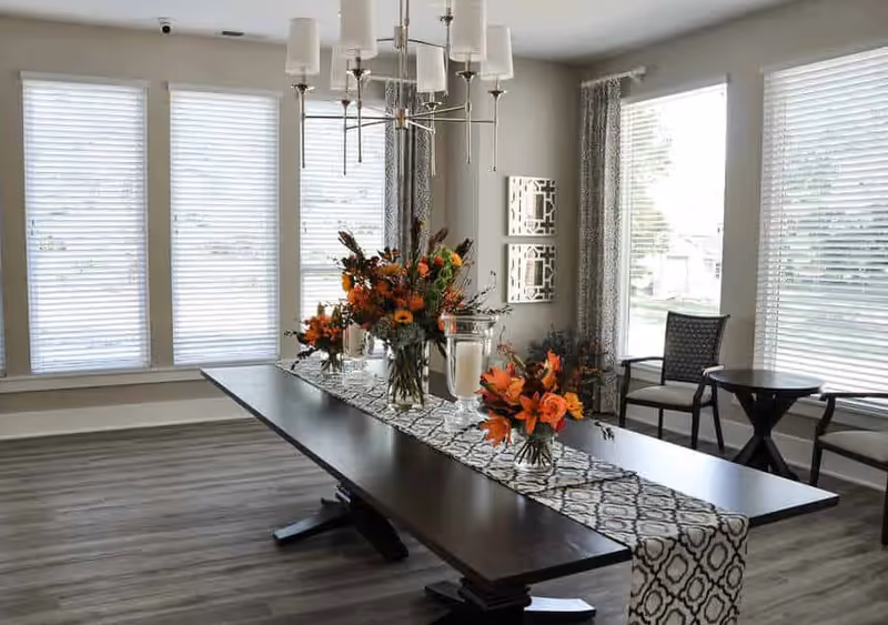 Sunlit dining room with a long dark wood table decorated with floral centerpieces and a chandelier, surrounded by large windows.