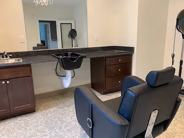 Interior view of a salon area in a senior living facility featuring a black salon chair facing a black hair washing sink attached to a dark granite countertop with wooden cabinets and drawers underneath. A large mirror is mounted on the wall behind the sink and chair. The floor has a patterned carpet and the walls are painted light beige.