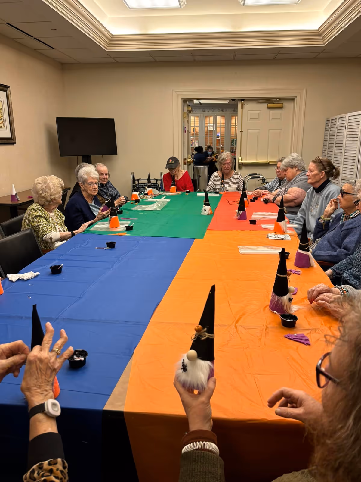 A group of elderly people sitting around a large table covered with colorful tablecloths in blue, green, and orange, engaging in a craft activity making small gnome decorations with black hats and white beards in a well-lit room with beige walls and a TV mounted on the wall.