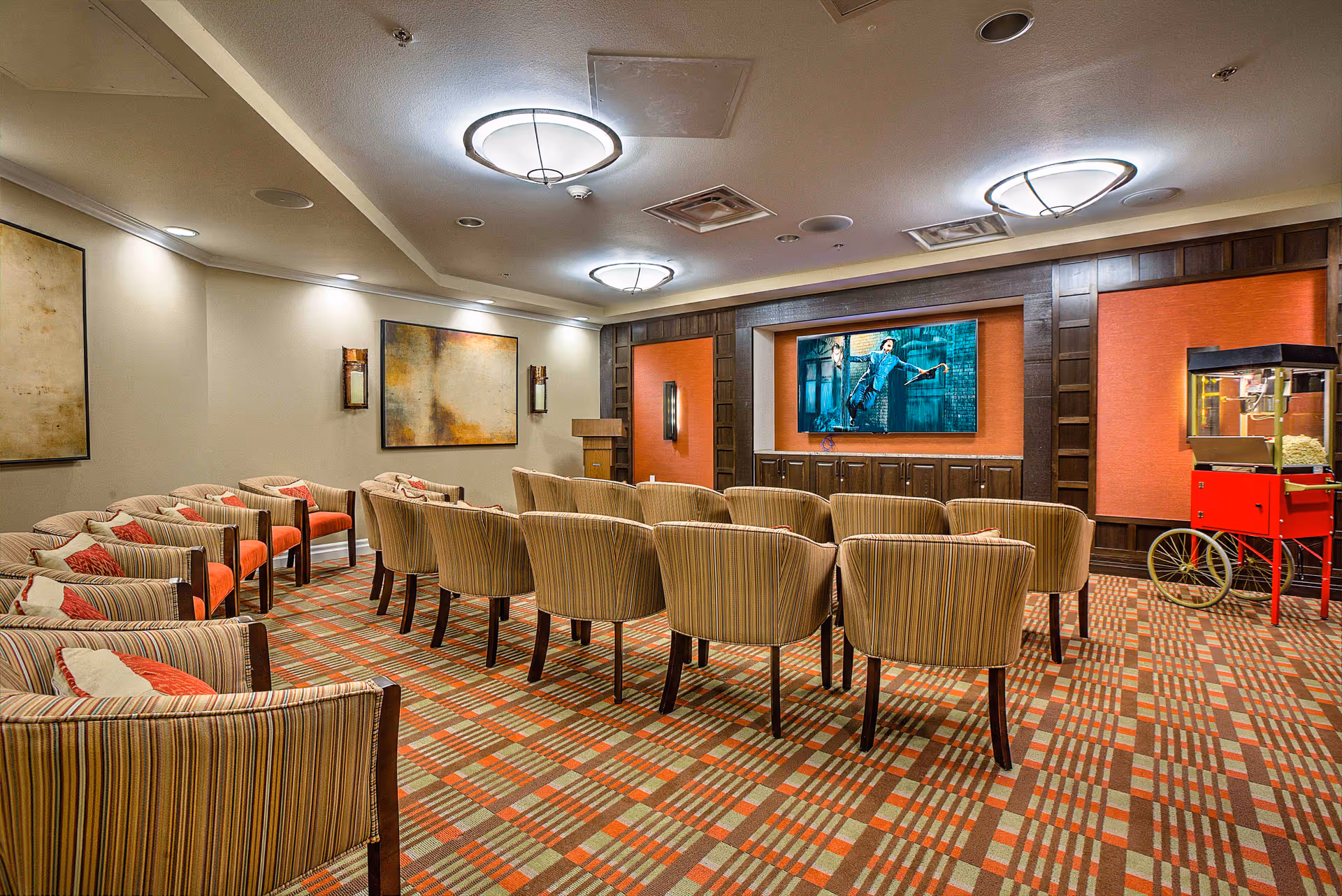 A cozy media room with multiple rows of striped upholstered chairs facing a large flat-screen TV mounted on a wall with orange panels and dark wood trim. The room has patterned carpet in shades of orange, beige, and brown, soft wall lighting, abstract artwork, and a vintage-style red popcorn machine on the right side.