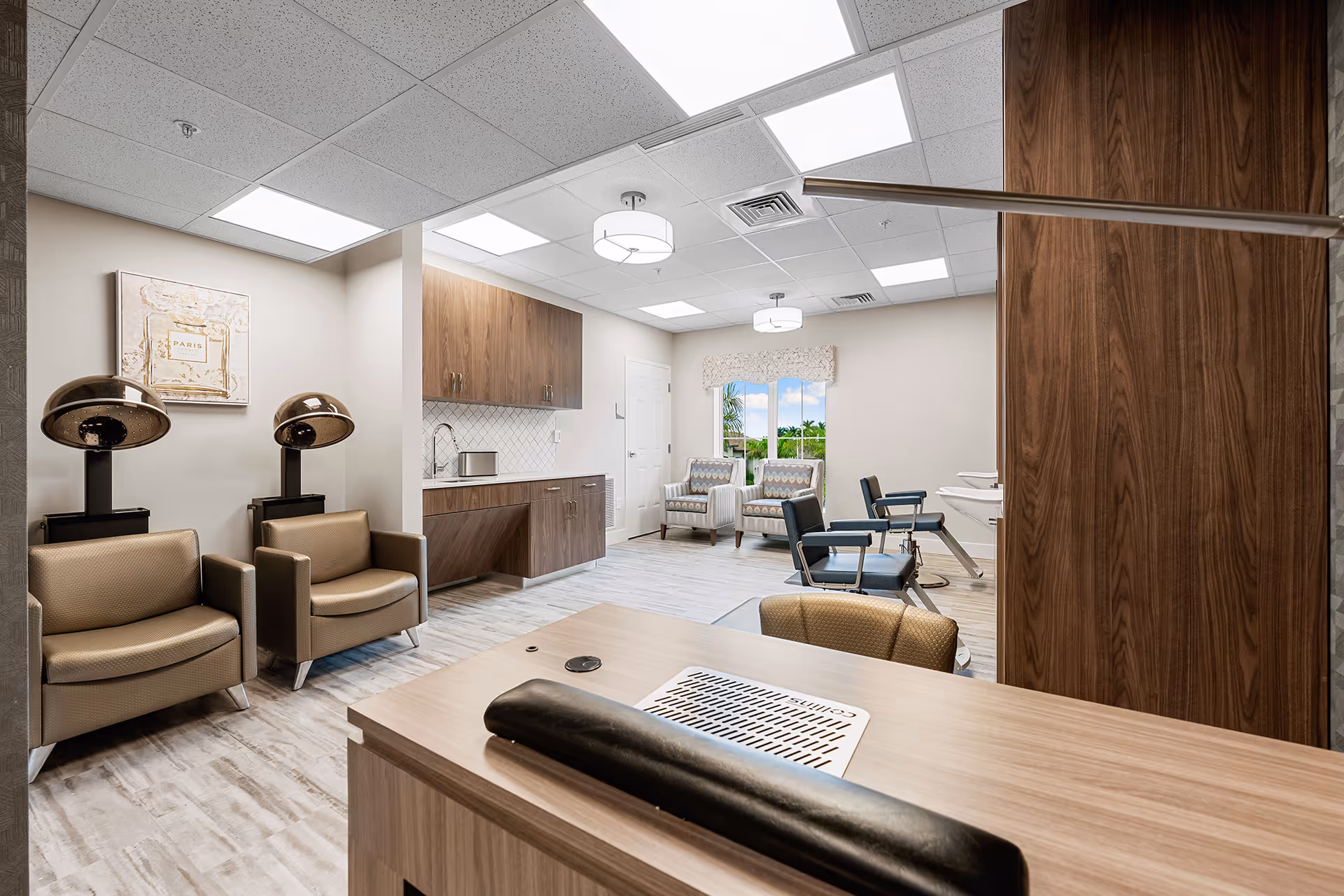 Interior view of a salon area in a senior living facility featuring two hair drying chairs with hooded dryers, two salon styling chairs with sinks, two armchairs near a window with a view of greenery outside, and wooden cabinetry along the wall under bright ceiling lights.