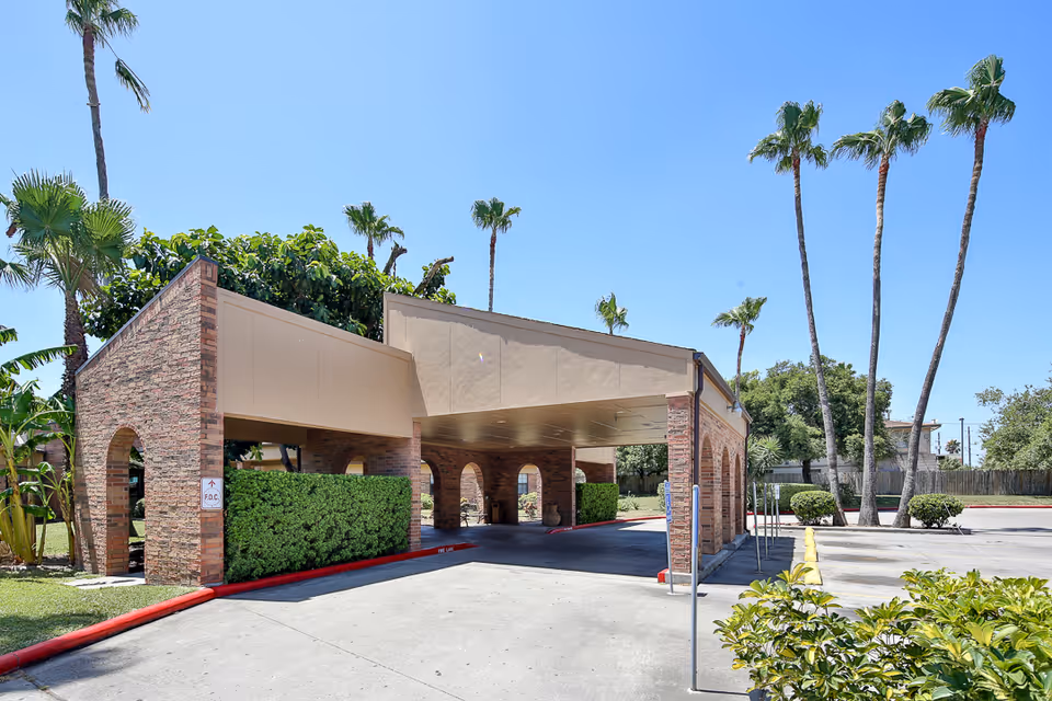 Covered driveway entrance to a building with brick pillars and arches, surrounded by palm trees and greenery under a clear blue sky.