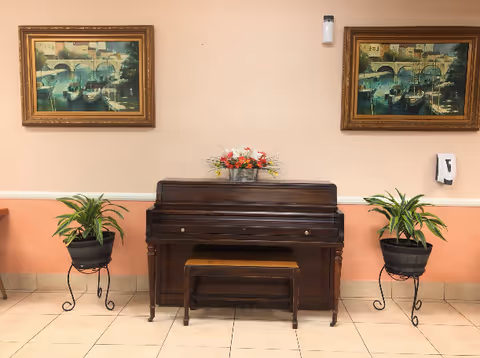 Upright piano with bench flanked by potted plants and two framed paintings on a peach-colored wall.