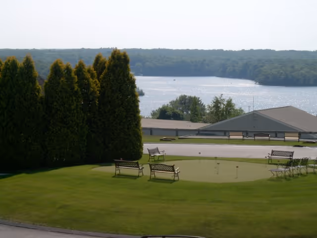 Well-maintained lawn with benches around a small putting green, a row of evergreen trees, and a lake visible beyond rooftops.
