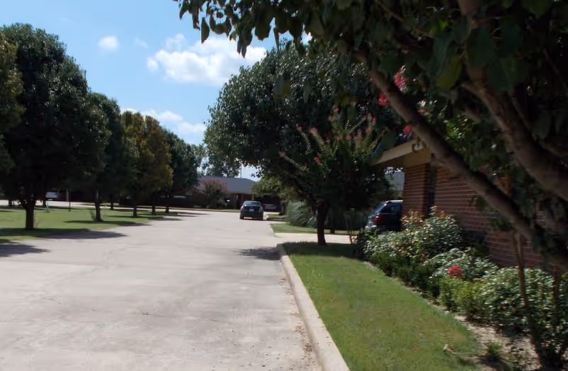A paved driveway lined with green grass and trees on both sides, with a brick building partially visible on the right. Two cars are parked along the driveway under a partly cloudy blue sky.