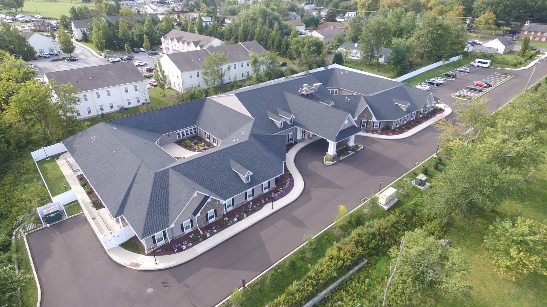 Aerial view of a large single-story building with a dark roof and white exterior walls, surrounded by landscaped greenery and parking areas. The building has a covered entrance and is situated in a suburban area with other buildings and trees nearby.