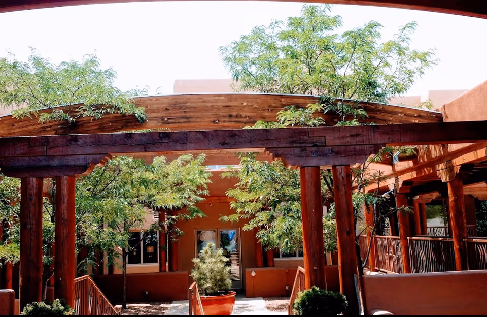 Wooden pergola-covered walkway leading to a courtyard entrance surrounded by trees and potted plants.