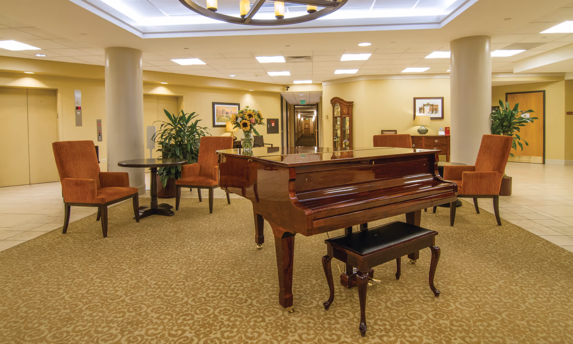 A spacious, well-lit common area featuring a polished grand piano with a matching bench on a patterned carpet. Surrounding the piano are four orange upholstered chairs and two small round tables. The background includes two elevators, potted plants, framed artwork, a wooden cabinet, and a hallway leading further into the building.