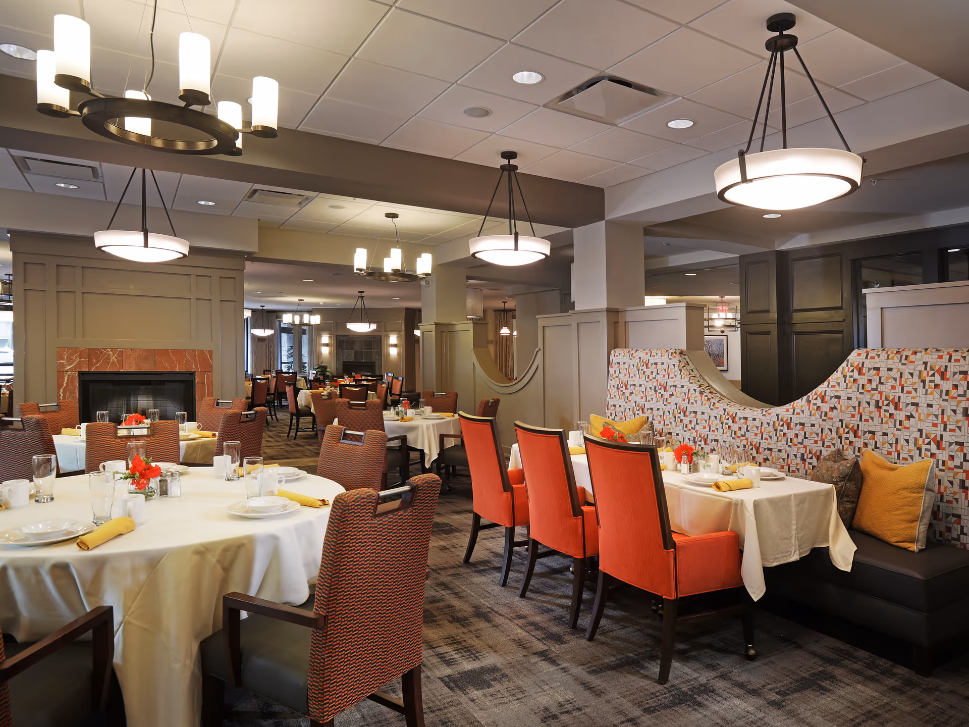 A spacious dining room with round and rectangular tables covered with white tablecloths, set with plates, glasses, and yellow napkins. The room features patterned chairs and booth seating with colorful cushions. Modern pendant lights hang from the ceiling, and a fireplace with a red marble surround is visible in the background.