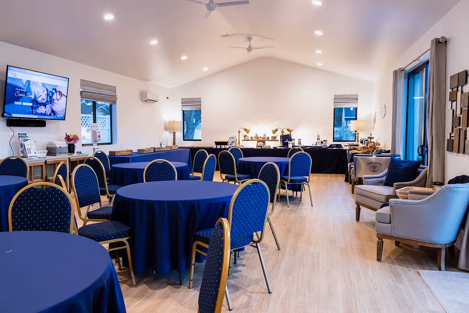 A well-lit community room with round tables covered in blue tablecloths and blue cushioned chairs arranged around them. The room has light wood flooring, white walls, and several windows with gray curtains. There is a TV mounted on the wall displaying a senior living website, a buffet table with lamps and various items at the back, and a seating area with two armchairs and a small table on the right side.
