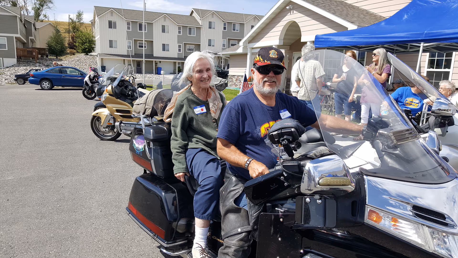 An elderly man and woman sitting on a black motorcycle in a parking lot outside a residential building. The man is wearing sunglasses, a black cap, and a blue t-shirt, while the woman is wearing a green sweater and blue pants. In the background, several other people are standing and sitting under a canopy near the building, with more motorcycles and cars parked nearby.