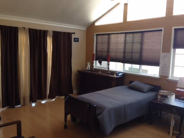 A bedroom in a senior living facility with a single bed covered in a gray bedspread and a matching pillow. There is a wooden dresser with decorative items and two red candles on it, positioned under a window with closed blinds. Dark brown curtains cover a door on the left side of the room. A small table with books and other items is next to the bed. The room has wooden flooring and beige walls.