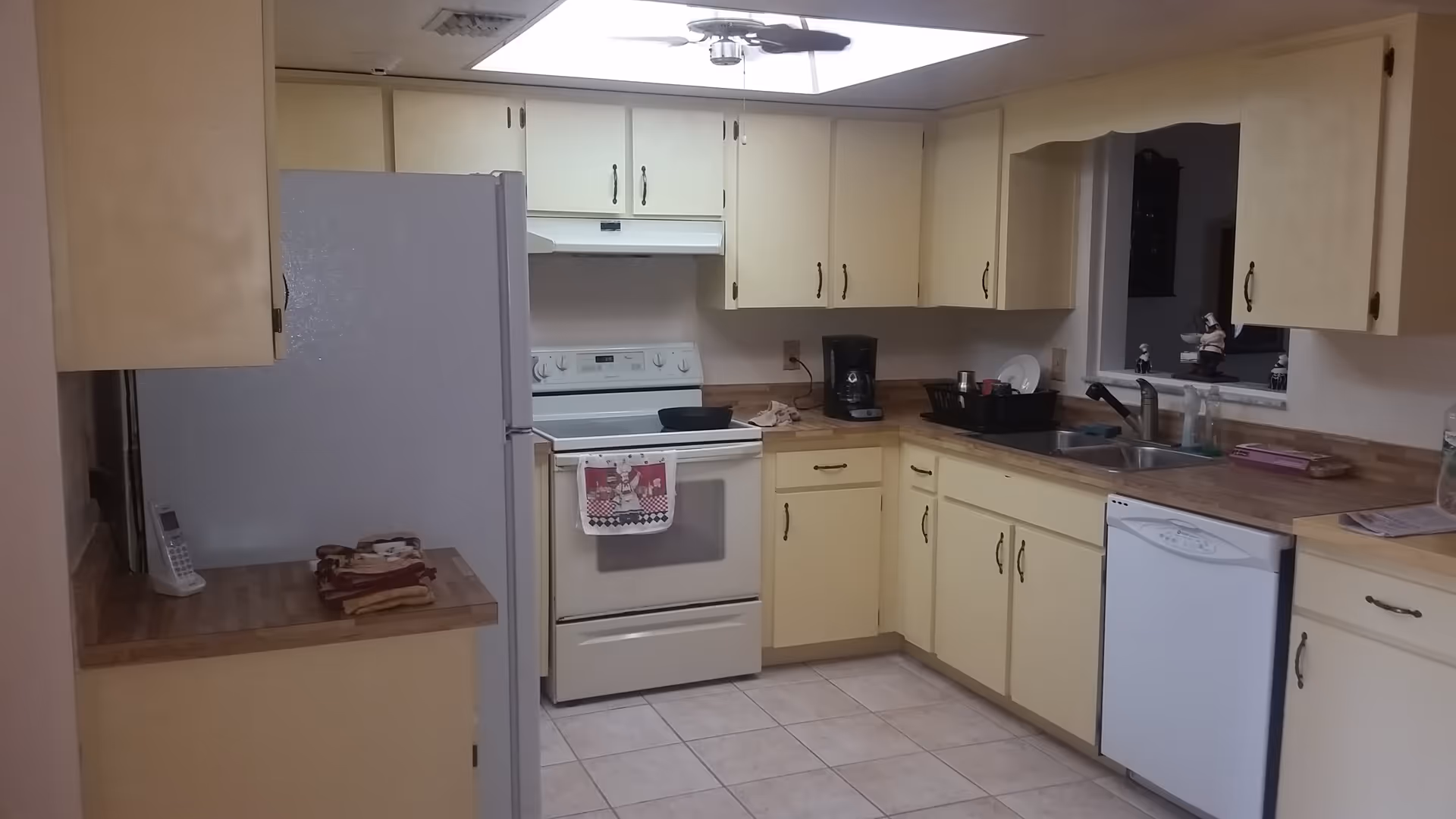 A kitchen with cream-colored cabinets and wood-patterned countertops. The kitchen includes a white refrigerator, a white electric stove with a towel hanging on the handle, a coffee maker, a double sink, and a white dishwasher. There are decorative figurines on the windowsill above the sink and a ceiling fan with a light fixture above.