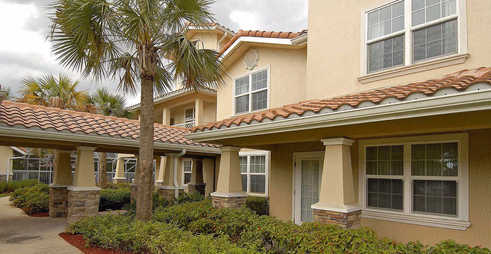 Exterior view of a beige two-story building with a tiled roof, surrounded by green shrubs and palm trees under a cloudy sky.