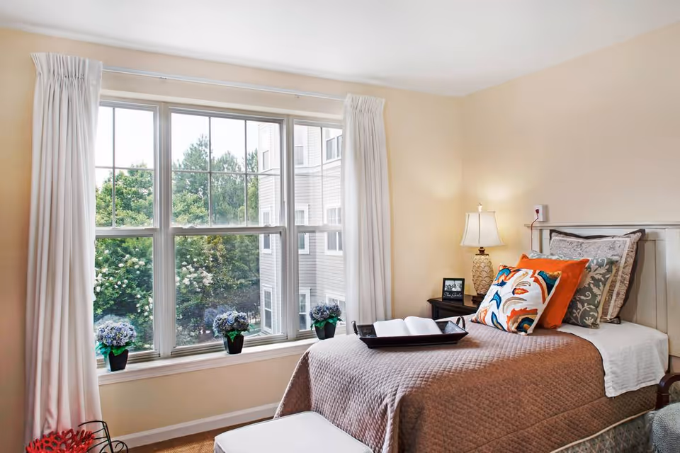 A cozy bedroom with a single bed covered in a brown quilt and multiple decorative pillows. A tray with an open book is placed on the bed. Next to the bed is a nightstand with a lamp and a small framed picture. The room has a large window with white curtains, showing green trees and part of a building outside. There are small potted plants on the windowsill.