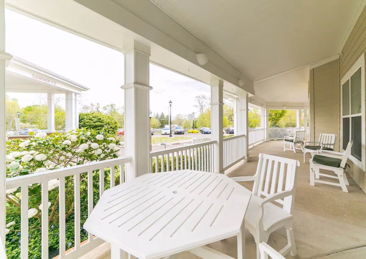Covered outdoor porch area with white railings and columns, featuring white patio furniture including a table and chairs. There are green bushes with white flowers along the railing and a parking lot with cars visible in the background.