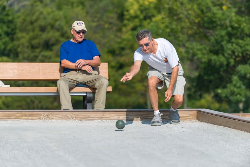 Two elderly men outdoors playing bocce ball on a sandy court. One man is sitting on a bench watching while the other is in the process of rolling a bocce ball. Both are casually dressed and wearing sunglasses, with greenery in the background.