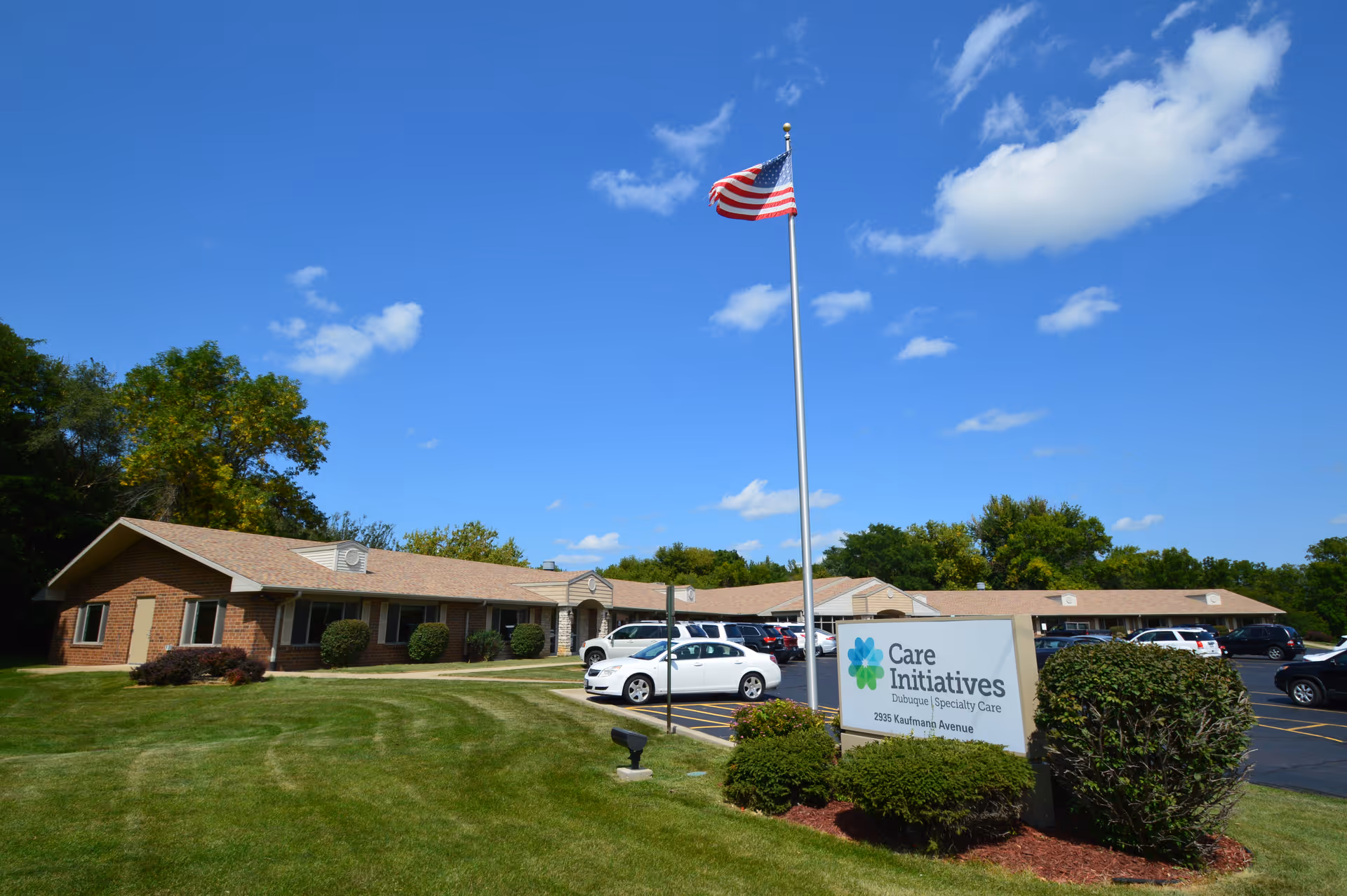 Exterior view of a single-story brick building with a parking lot in front, an American flag on a tall flagpole, and a sign that reads 'Care Initiatives Dubuque Specialty Care 2355 Kaufmann Avenue' surrounded by green bushes and a well-maintained lawn under a blue sky with scattered clouds.